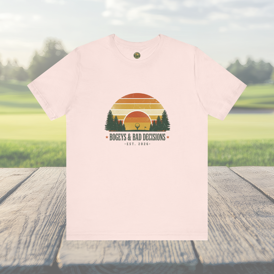 Pink t-shirt with a sunset graphic and text on a wooden surface with a grassy field background